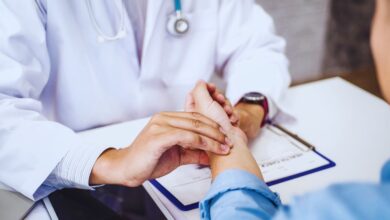 Image shows A doctor in a white coat gently examining a patient's hand during a medical consultation.