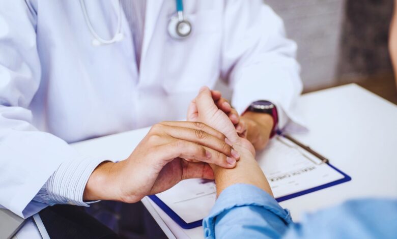 Image shows A doctor in a white coat gently examining a patient's hand during a medical consultation.