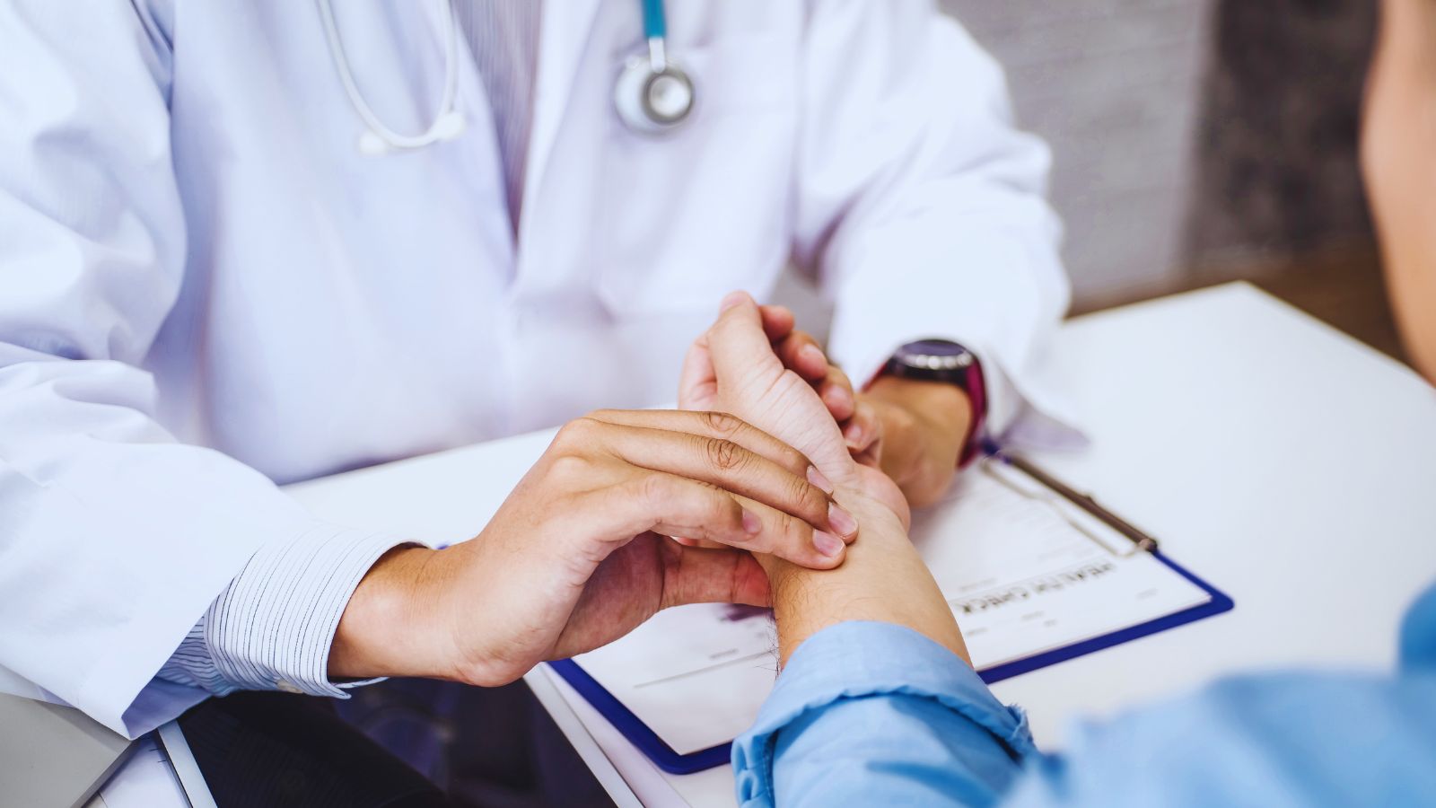 Image shows A doctor in a white coat gently examining a patient's hand during a medical consultation.