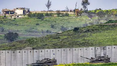 Two tanks positioned near a border wall with a hilly landscape and damaged buildings in the background.