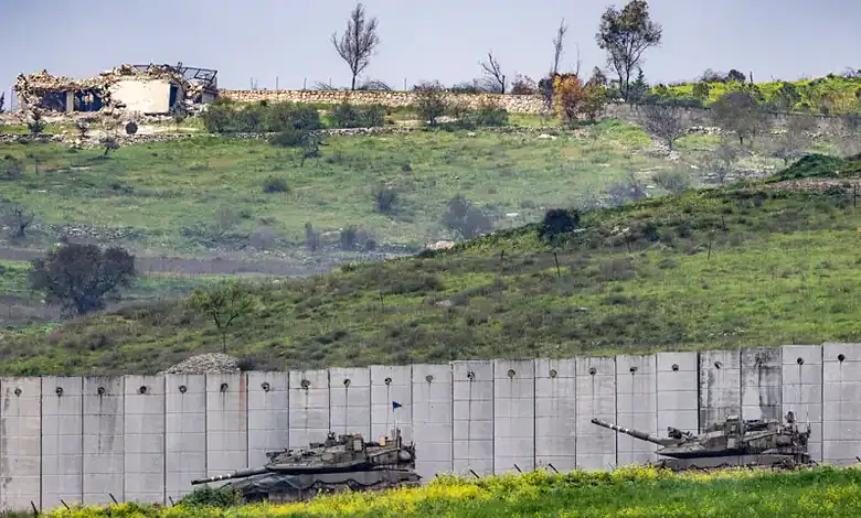 Two tanks positioned near a border wall with a hilly landscape and damaged buildings in the background.