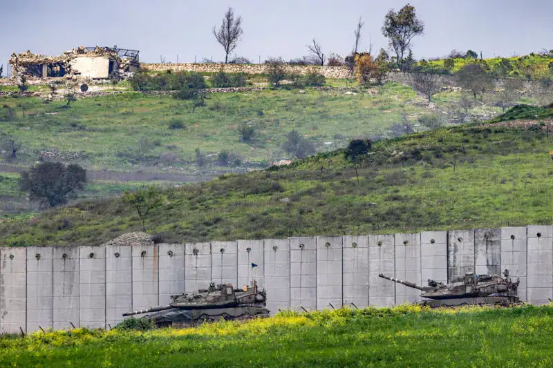 Two tanks positioned near a border wall with a hilly landscape and damaged buildings in the background.
