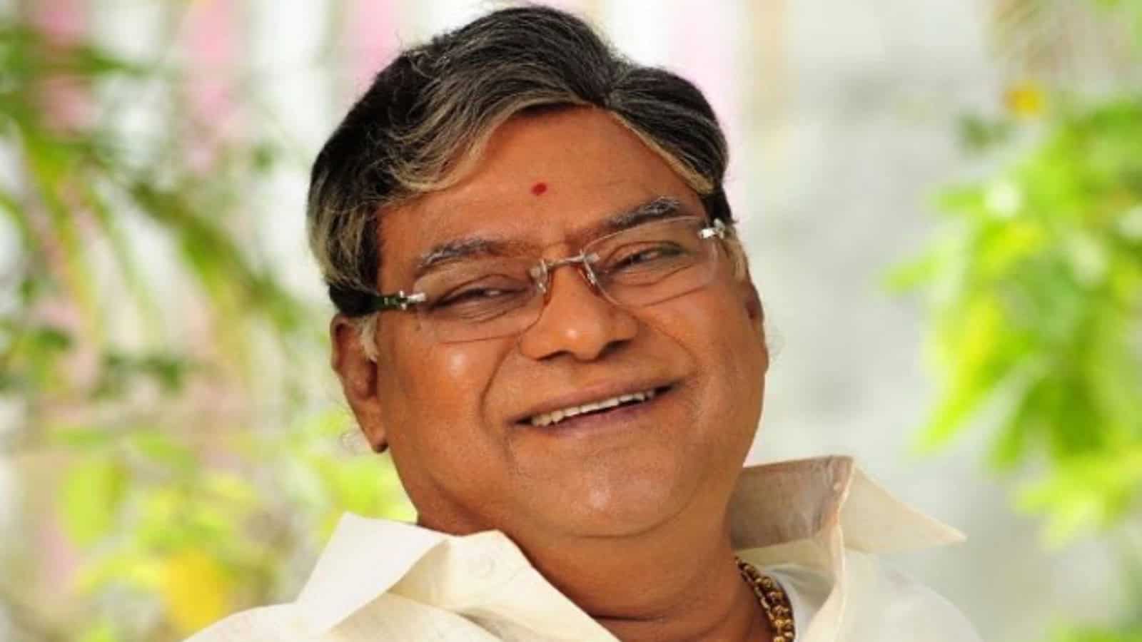 An elderly Indian man smiling warmly, wearing traditional attire and glasses, in a bright, natural settin.