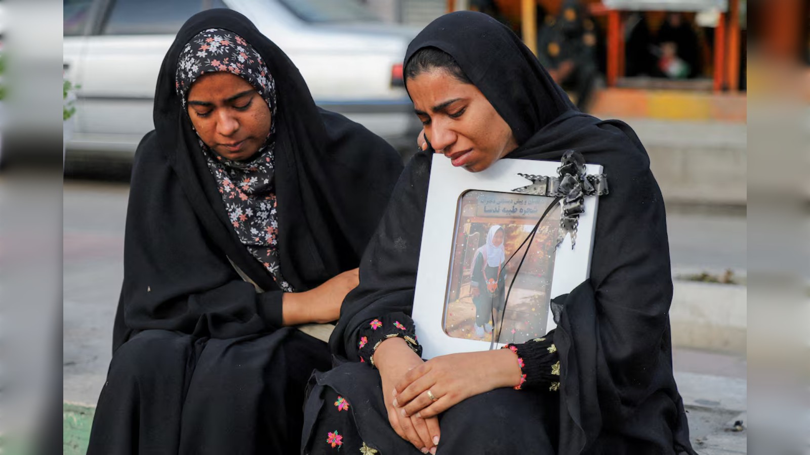 Two Iranian women mourning, holding a framed photo of schoolgirls, reflecting tragedy and loss.