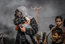 A Palestinian woman holds a child as smoke rises in the background in Gaza while other children stand nearby.