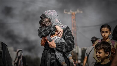 A Palestinian woman holds a child as smoke rises in the background in Gaza while other children stand nearby.