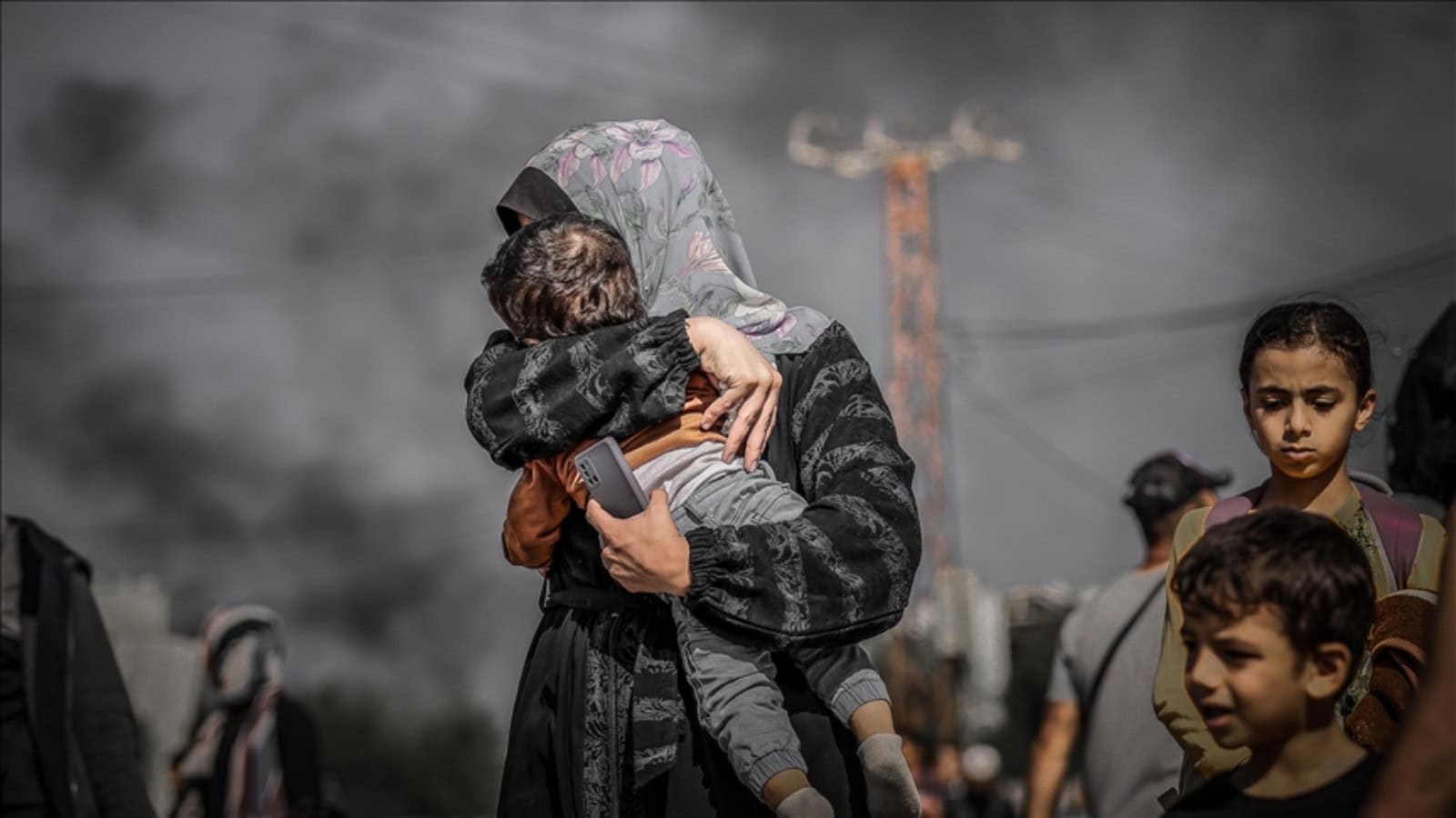A Palestinian woman holds a child as smoke rises in the background in Gaza while other children stand nearby.