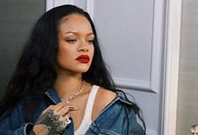 Rihanna with long black hair and red lipstick, wearing a denim jacket, in a home interior.