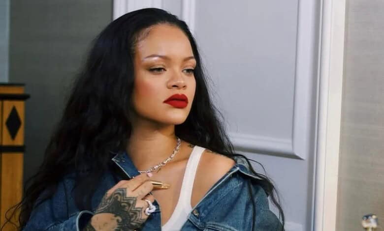 Rihanna with long black hair and red lipstick, wearing a denim jacket, in a home interior.