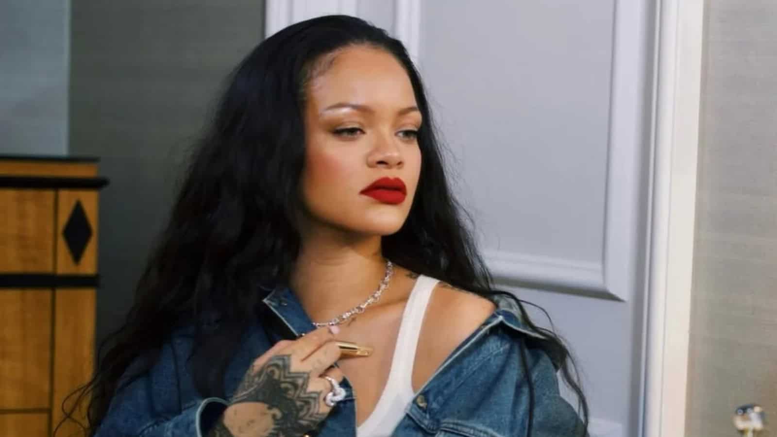 Rihanna with long black hair and red lipstick, wearing a denim jacket, in a home interior.