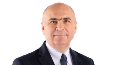 A professional portrait of a Romanian politician in a dark suit and tie, smiling confidently against a pl.