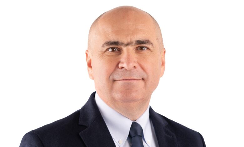 A professional portrait of a Romanian politician in a dark suit and tie, smiling confidently against a pl.