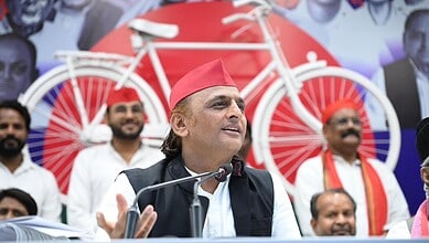 Akhilesh Yadav speaking at a rally with supporters and red bicycle props in the background.