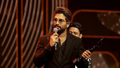 Allu Arjun speaking into a microphone during a film shoot in Mumbai, holding sunglasses, with a warm back.