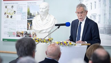 Austrian president speaking at a podium during a public event, with a bust and informational posters in t.