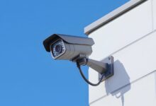 Image shows A wall-mounted CCTV security camera with infrared LEDs is affixed to the corner of a white building against a clear blue sky.