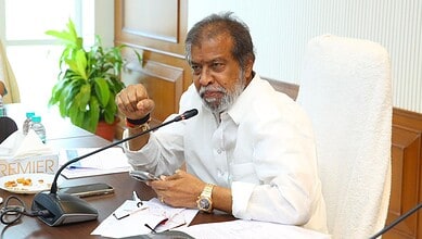 An elderly man speaking at a meeting about Telangana's public healthcare system reform, seated at a desk.