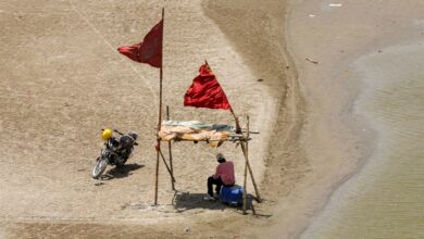 Small makeshift shelter on a sandy beach with a person sitting under it, near water, during a heatwave.