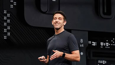 An enthusiastic male speaker presenting at a tech conference, holding a remote control, with a digital ba.