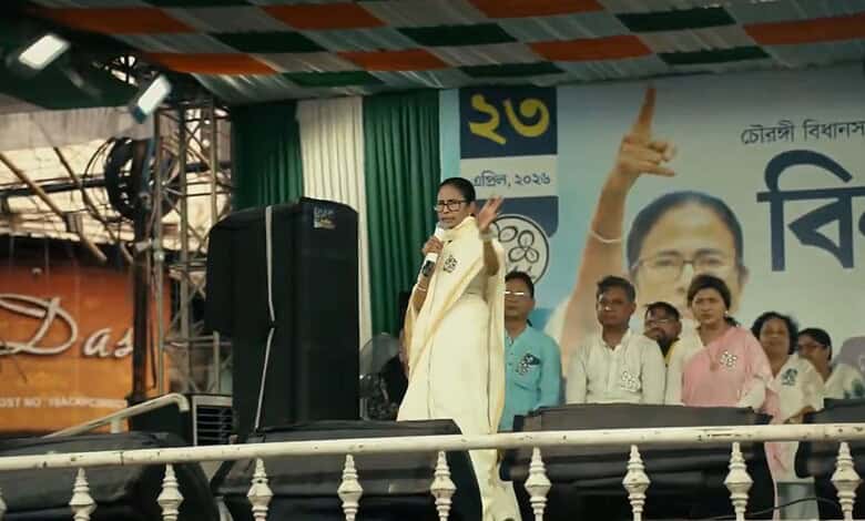 Politician speaking at a public rally with supporters and a large poster in the background.