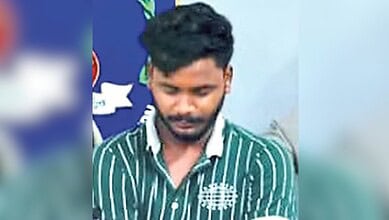 Young man with black hair and beard wearing a green striped shirt, looking down, in an indoor setting.