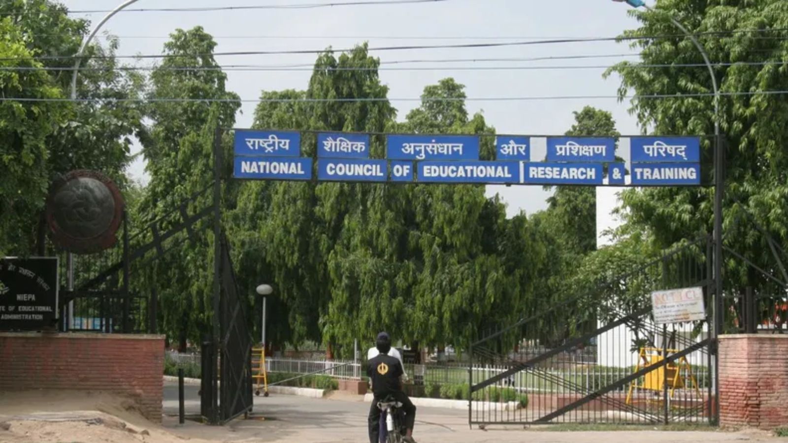Image shows NCERT headquarters entrance gate with blue signboard displaying the organization's name in Hindi and English.