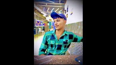 Young man in a blue cap and checkered shirt sitting in a public space, possibly Talabkatta, Hyderabad.