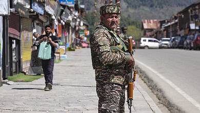 Soldier in camouflage uniform with rifle on a street in a mountainous area, with shops and pedestrians in.