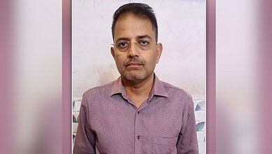 Man with glasses wearing a casual shirt, standing indoors against a plain background.