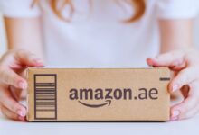 A person holding a cardboard Amazon.ae delivery package with a barcode on a white background.