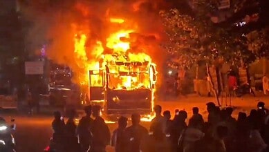Bus engulfed in flames with onlookers watching nearby.