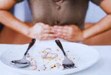 Image shows A person clutching their stomach in discomfort behind a plate of leftover seafood scraps, illustrating food poisoning or digestive distress.