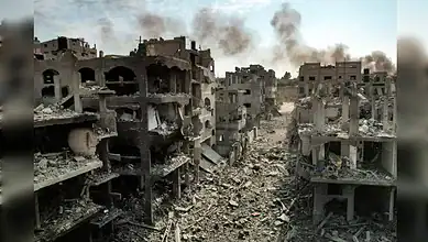 Ruined buildings and debris in a conflict zone, with smoke rising in the background.