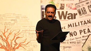Indian man speaking at a press conference with newspaper headlines in the background.