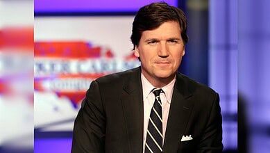 Tucker Carlson poses in a television studio wearing a dark suit and striped tie.
