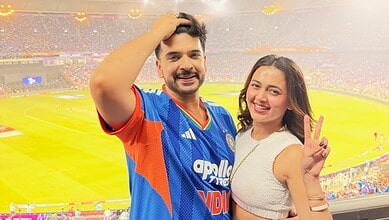 Tejasswi Prakash smiling and making a peace sign at a cricket stadium.