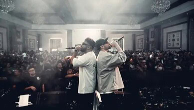 Two hip-hop artists performing on stage at 'LEGACY' show with a large audience in the background.