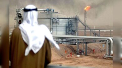 Oil facility in desert with gas flare burning as a man in traditional Gulf attire looks on.