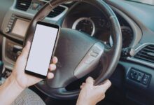 Image shows A driver holding a smartphone with a blank screen while steering a car with their other hand.