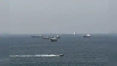 Cargo ships anchored in the Strait of Hormuz with a speedboat passing during shipping disruption.