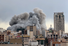 Thick grey smoke billows over buildings in Tehran after an air strike, rising above the urban skyline.