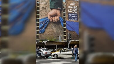 Hand gripping blue fabric in front of a large billboard in Iran, with cars and pedestrians nearby.