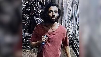 A man with long hair and a beard, dressed in a red shirt, standing outside near a metal fence following an incident involving security guards in Maharashtra.