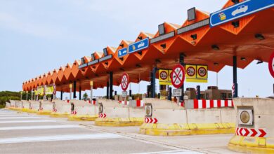 Image shows A multi-lane highway toll plaza with a distinctive orange zigzag canopy roof, traffic signs, and concrete lane dividers.