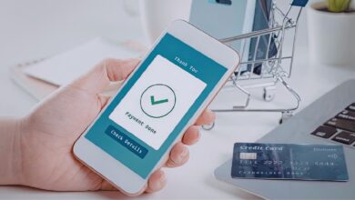 Image shows A hand holds a smartphone showing a "Payment Done" confirmation screen, with a credit card and miniature shopping cart on a desk nearby.