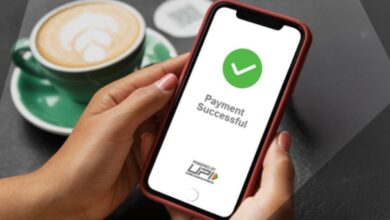 Image shows A hand holds a smartphone displaying a "Payment Successful" confirmation screen powered by UPI, with a coffee cup in the background.