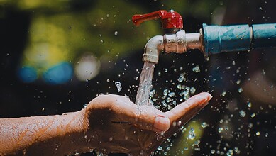 Close-up of a hand catching water from a leaking outdoor tap with blurred greenery background.