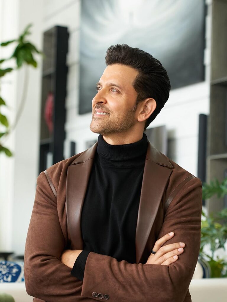 A confident man with dark hair and beard, wearing a black turtleneck and brown blazer, smiling thoughtful.