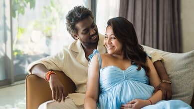 Happy couple sitting on sofa, smiling, with pregnant woman in blue dress, celebrating pregnancy indoors.