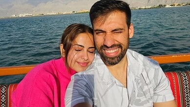 A smiling couple taking a selfie on a boat with water and mountains in the background.